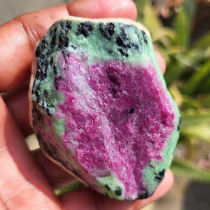 May include: A raw gemstone with a striking magenta core, framed by a green border and dotted with black specks. The stone's natural, unrefined texture and colour variations are visible, held in a hand.