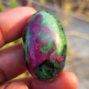 Może przedstawiać: Owalny kamień szlachetny o wypolerowanej powierzchni. Kamień ma mieszankę kolorów, w tym głęboką czerwień, zieleń i czerń. Kamień szlachetny jest trzymany między palcami, prezentując jego naturalne piękno i unikalne wzory kolorystyczne. Idealny do tworzenia biżuterii.