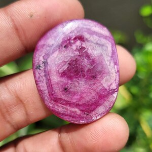 May include: A polished, oval gemstone in a deep magenta colour, with a mix of lighter and darker shades. The stone has a translucent quality, with visible internal patterns and inclusions. The surface is smooth and reflects light, showcasing its natural beauty.