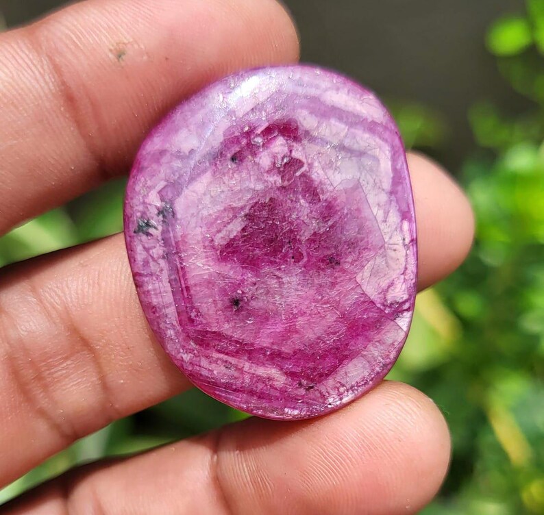 May include: A polished, oval gemstone in a deep magenta colour, with a mix of lighter and darker shades. The stone has a translucent quality, with visible internal patterns and inclusions. The surface is smooth and reflects light, showcasing its natural beauty.