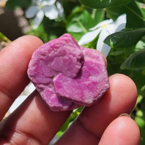 May include: A raw, hexagonal-shaped ruby crystal with a vibrant magenta-pink hue. The stone features a rough, natural texture with visible crystalline layers and geometric facets, showcasing a deep, saturated color throughout its entire structure.