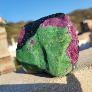 May include: A rough-cut natural stone with a mix of colors. The stone features a vibrant green base with patches of deep magenta and black. The stone is sitting on a light gray surface.