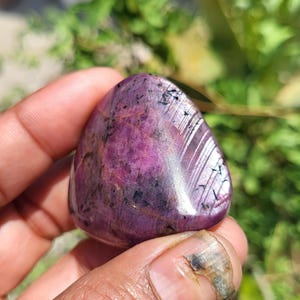 Puede incluir: Una gema pulida, de forma ovalada, con un tono magenta intenso, salpicada de inclusiones negras. La piedra tiene una superficie lisa y reflectante, con vetas de color más claro. La gema se sostiene en una mano, con un fondo verde borroso.