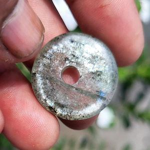 Könnte beinhalten: Ein kreisförmiger, durchscheinender Steinanhänger mit polierter Oberfläche, der ein zentrales Loch und interne Mineraleinschlüsse aufweist, die ein schimmerndes, mehrfarbiges Aussehen mit silbernen, grauen Flecken und subtilen irisierenden Reflexen erzeugen.