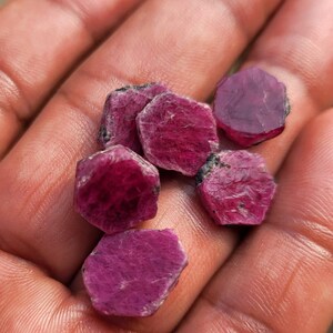 May include: A handful of raw, uncut ruby gemstones. The gemstones are a deep, rich red color with a slightly rough, textured surface. The rubies are hexagonal in shape and vary slightly in size.