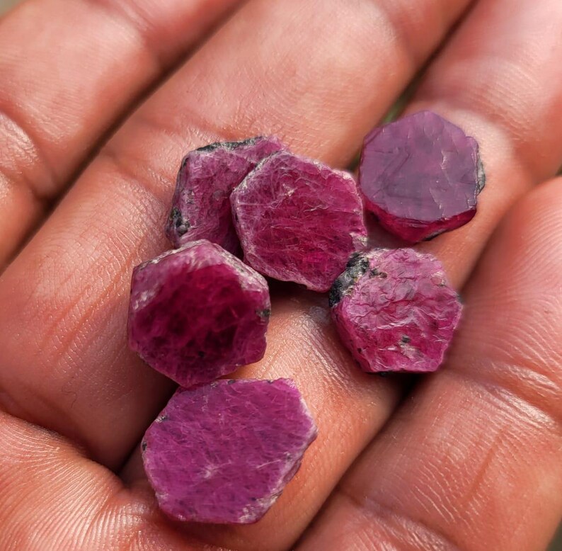 May include: A handful of raw, uncut ruby gemstones. The gemstones are a deep, rich red color with a slightly rough, textured surface. The rubies are hexagonal in shape and vary slightly in size.