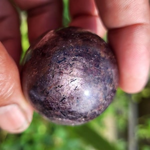 Peut inclure: Gros plan sur une pierre précieuse sphérique polie. La pierre a une teinte violet foncé avec des inclusions chatoyantes et irisées et une surface texturée. La sphère est tenue entre les doigts sur un fond vert flou.