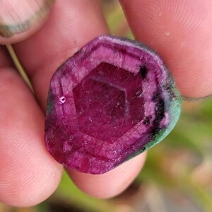 May include: A faceted, oval gemstone with a deep magenta centre and a dark green outer edge. The gem's interior features a geometric pattern, held between fingers.