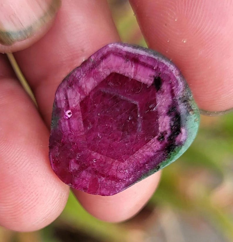 May include: A faceted, oval gemstone with a deep magenta centre and a dark green outer edge. The gem's interior features a geometric pattern, held between fingers.