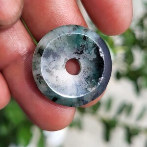 May include: A circular stone pendant featuring a polished surface with a central hole. The stone displays a mottled pattern of translucent white, deep forest green, and dark charcoal gray inclusions throughout its smooth, rounded form.