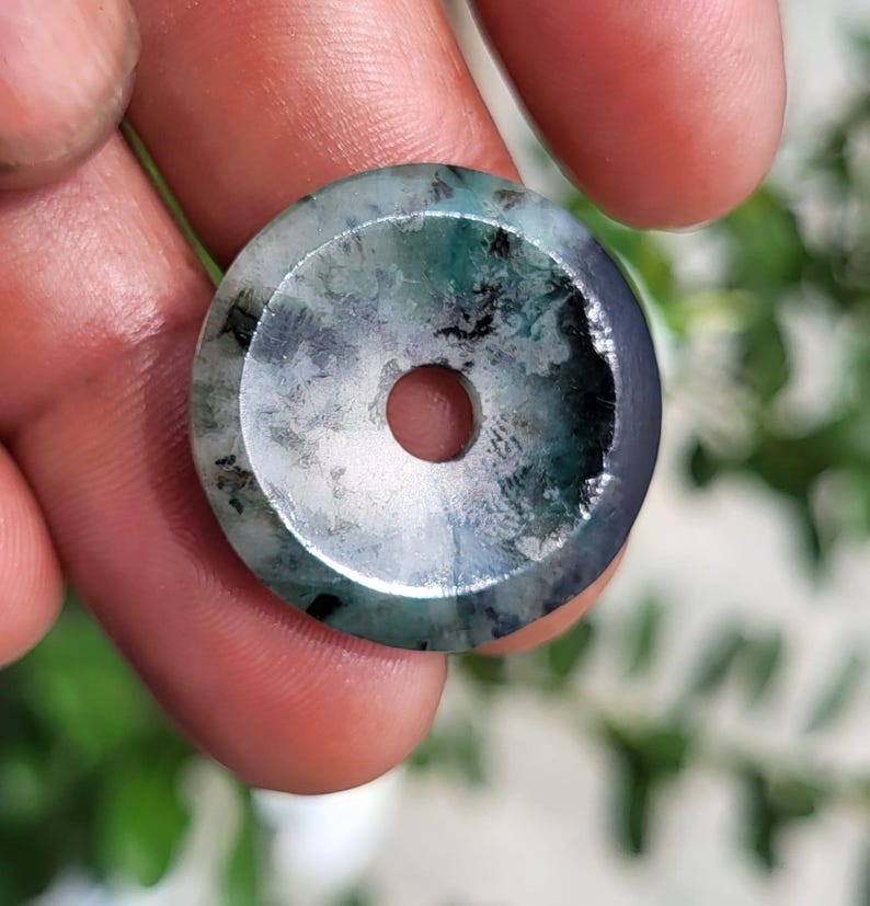 May include: A circular stone pendant featuring a polished surface with a central hole. The stone displays a mottled pattern of translucent white, deep forest green, and dark charcoal gray inclusions throughout its smooth, rounded form.
