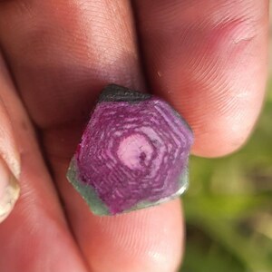 Pode incluir: Um close-up de uma pedra preciosa segurada em uma m&atilde;o. A pedra tem um padr&atilde;o conc&ecirc;ntrico em camadas em tons de rosa e roxo, com uma borda verde e preta. A superf&iacute;cie da pedra &eacute; &aacute;spera e natural.