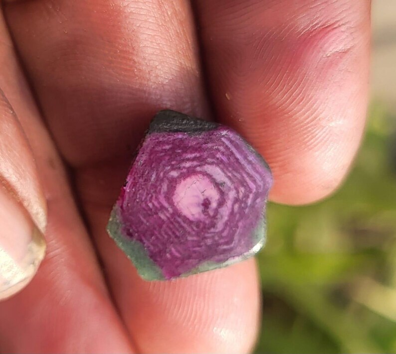 Pode incluir: Um close-up de uma pedra preciosa segurada em uma m&atilde;o. A pedra tem um padr&atilde;o conc&ecirc;ntrico em camadas em tons de rosa e roxo, com uma borda verde e preta. A superf&iacute;cie da pedra &eacute; &aacute;spera e natural.