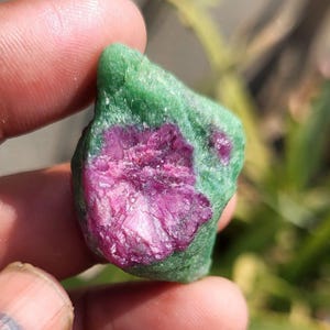Könnte beinhalten: Ein naturbelassener Edelstein mit einem auffälligen Farbkontrast. Die Außenseite ist tiefgrün, während das Innere ein lebendiges Fuchsia-Rosa aufweist. Der grob geschnittene Stein wird in einer Hand gehalten, was sein rohes, unpoliertes Aussehen hervorhebt.