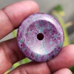 Può includere: Un ciondolo rotondo in pietra preziosa lucidata con un foro centrale. La pietra presenta una miscela di tonalità rosa intenso e verde, con una finitura liscia e lucida. Il ciondolo è tenuto in una mano.