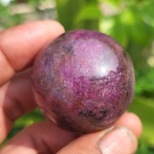 Op de afbeelding: Een gepolijste, bolvormige robijn met een diepe magenta en paarse tint, voorzien van zichtbare interne kristalstructuren, donkere minerale insluitingen en een glad, reflecterend oppervlak dat het licht opvangt.