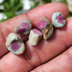 Puede incluir: Cinco piedras preciosas pequeñas, en bruto y sin pulir, con centros de color magenta vibrante incrustados en una matriz mineral rugosa de color verde menta pálido. Cada piedra tiene una forma orgánica irregular con una superficie texturizada, mostrando un contraste natural entre el núcleo rosa intenso y la capa exterior verde terrosa más clara.
