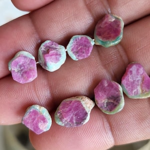 Peut inclure: Un ensemble de perles de pierres précieuses de rubis brutes. Les perles sont d'un rouge-rosé vif avec une matrice bleu-vert clair. Les pierres précieuses sont de forme irrégulière et enfilées, mettant en valeur leur beauté naturelle.