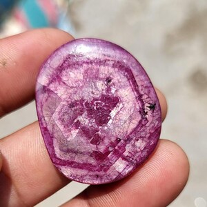 May include: A polished, oval gemstone in a deep magenta colour. The stone displays a complex internal pattern of concentric circles and geometric shapes, with darker veins and inclusions. The surface is smooth and reflective, giving it a glossy appearance.