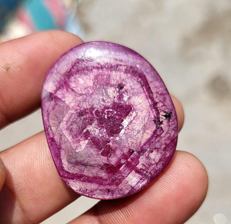 May include: A polished, oval gemstone in a deep magenta colour. The stone displays a complex internal pattern of concentric circles and geometric shapes, with darker veins and inclusions. The surface is smooth and reflective, giving it a glossy appearance.