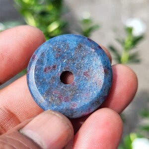 May include: A circular, flat, blue stone pendant with a central hole. The surface features a mottled texture with deep blue tones and scattered dark red and brown inclusions throughout the polished, rounded stone disc.