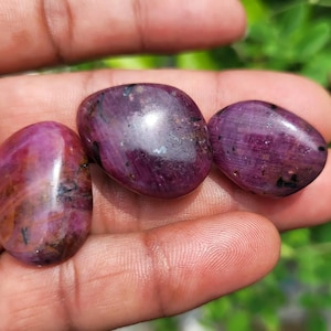 Può includere: Tre pietre preziose lucide, di forma ovale, in varie tonalità di rosso intenso e viola. Le pietre hanno una superficie liscia e riflettente con sottili variazioni di colore e consistenza. Le pietre sono tenute in una mano.