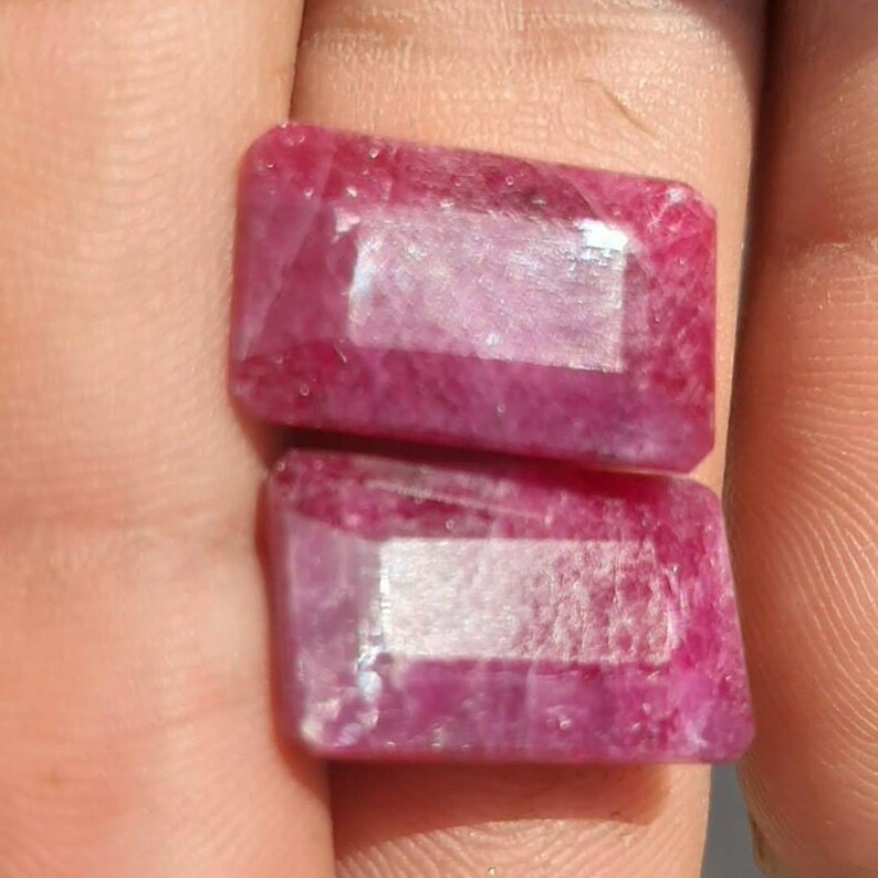 May include: Two rectangular, faceted ruby gemstones. The gems are a deep, saturated pinkish-red colour with internal inclusions. The facets catch the light, creating a sparkling effect. These gemstones are likely for jewellery making.