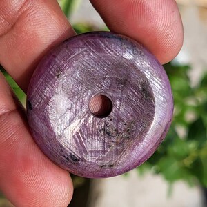 May include: A circular, flat, purple-toned ruby gemstone pendant with a polished surface and a central hole. The stone features a distinct cross-hatched, fibrous texture throughout its surface, with darker inclusions and a translucent quality.