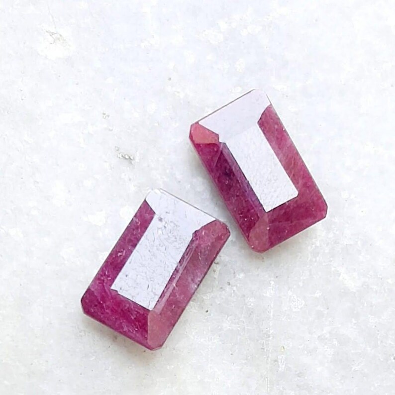 May include: Two rectangular, faceted gemstones in a deep pink colour. Each gem features a white, rectangular area in the centre, contrasting with the pink. The gems are set against a white, textured background.