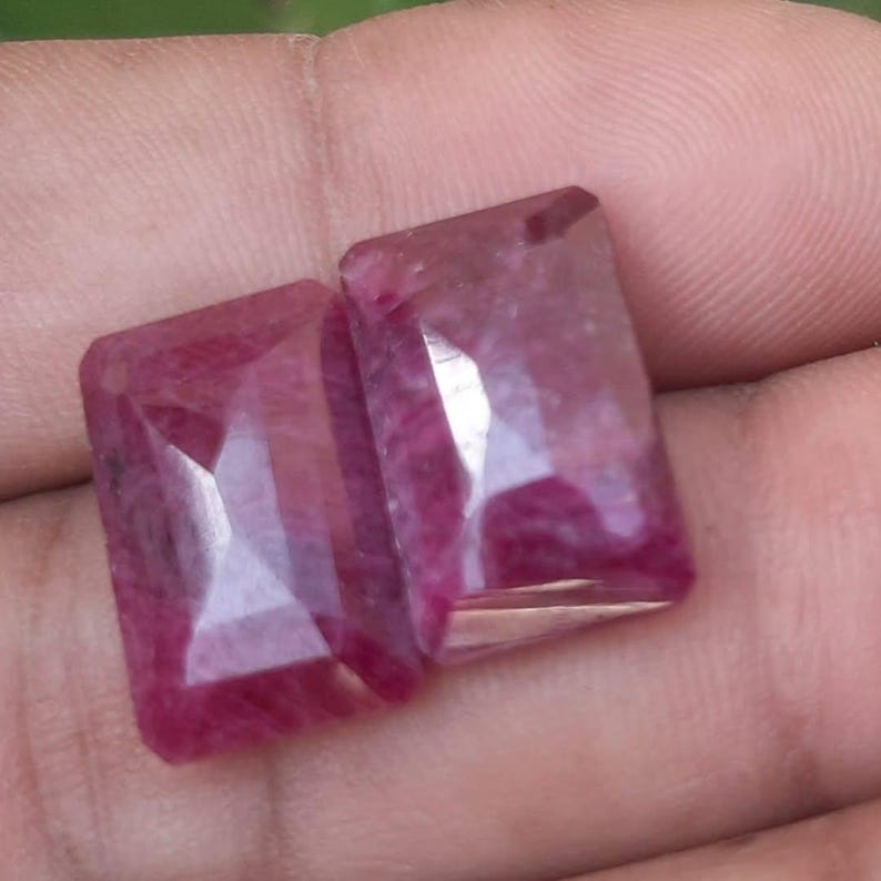 May include: Two faceted, rectangular ruby gemstones. The gems are a deep, rich red colour with internal inclusions. The cut of the stones creates multiple reflective surfaces, enhancing their brilliance. The gems are held in a hand.