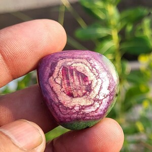 May include: A polished, rounded gemstone featuring a deep magenta and purple centre with a hexagonal, light-coloured crystalline pattern. The outer edges of the stone transition into a dark green hue, creating a distinct contrast in colour and texture.