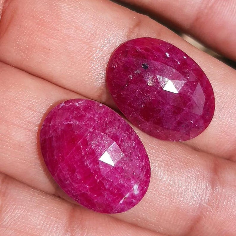 May include: Two oval-shaped, faceted ruby gemstones. The rubies are a deep, vibrant red color, with multiple facets that catch the light. The gemstones are displayed on a light-colored surface.