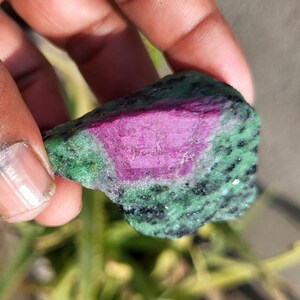 Op de afbeelding: Een ruwe edelsteen met een levendig roze kern omgeven door een gespikkelde groene buitenkant. De steen heeft een natuurlijke, onregelmatige textuur, met een mix van kleuren en patronen. De edelsteen wordt in een hand gehouden.