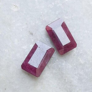 May include: Two rectangular, faceted gemstones in a deep red colour. Each gem has a lighter, rectangular interior, creating a striking contrast. The gems are displayed on a light, textured surface, highlighting their colour and cut. Suitable for jewellery making.