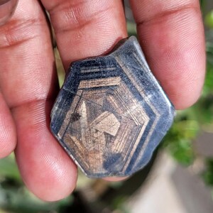 May include: A dark, geometric gemstone with a complex internal pattern. The stone has a hexagonal shape with a dark blue-black exterior and a lighter, tan-colored interior. The surface has visible lines and imperfections.