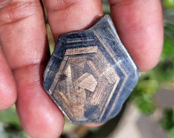 May include: A dark, geometric gemstone with a complex internal pattern. The stone has a hexagonal shape with a dark blue-black exterior and a lighter, tan-coloured interior. The surface has visible lines and imperfections.