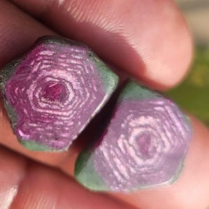 Pode incluir: Duas pedras preciosas em bruto, com um tom magenta profundo e um padr&atilde;o &uacute;nico em forma de estrela. As pedras t&ecirc;m uma borda verde e s&atilde;o seguradas em uma m&atilde;o, mostrando sua beleza natural e textura.