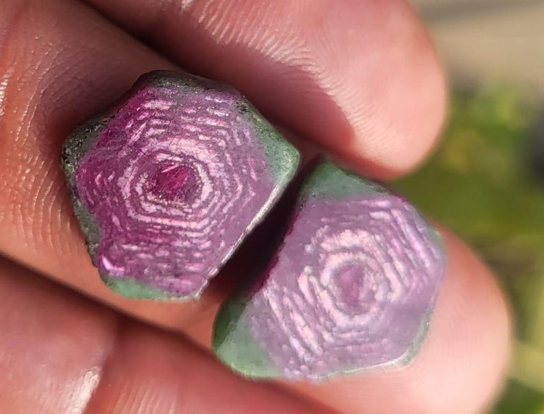 Pode incluir: Duas pedras preciosas em bruto, com um tom magenta profundo e um padr&atilde;o &uacute;nico em forma de estrela. As pedras t&ecirc;m uma borda verde e s&atilde;o seguradas em uma m&atilde;o, mostrando sua beleza natural e textura.