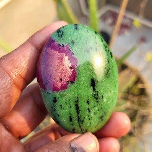 May include: An oval-shaped gemstone with a vibrant green base, speckled with black and featuring a prominent patch of deep magenta. The polished stone has a smooth, glossy surface.