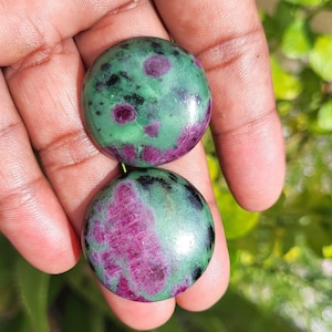 Può includere: Due pietre preziose rotonde e lucide. Le pietre presentano un mix di verde e nero con inclusioni color magenta vibrante. Le pietre preziose sono lisce e sembrano naturali, con una finitura lucida. Le pietre sono tenute in una mano.