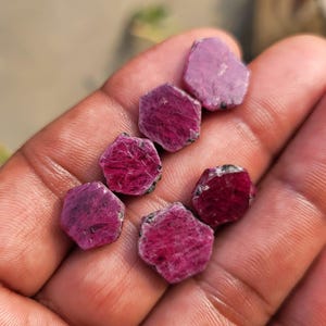 Op de afbeelding: Een verzameling ruwe robijn edelstenen in verschillende tinten roze en rood. De ongeslepen stenen hebben een natuurlijk, getextureerd uiterlijk, met enkele donkergrijze of zwarte insluitsels. De edelstenen worden in een hand gehouden.