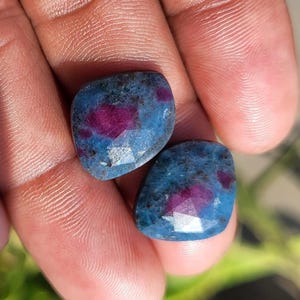May include: Two polished gemstones with a blue and red color scheme. The stones have a faceted surface and a mix of blue and red hues. The gemstones are held in a hand, with a blurred green background.