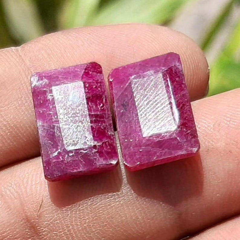 May include: Two rectangular, faceted ruby gemstones. Each gem has a deep pinkish-red hue with a lighter, rectangular, reflective area in the centre. The gems are held between fingers, with a blurred green background.