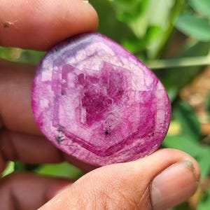 May include: A polished, oval gemstone in a deep magenta colour. The stone has a translucent quality, with visible internal patterns and inclusions. The gem is held between fingers, with a blurred green background.