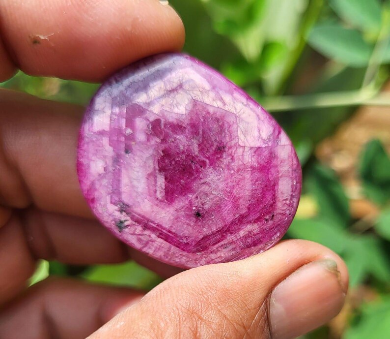 May include: A polished, oval gemstone in a deep magenta colour. The stone has a translucent quality, with visible internal patterns and inclusions. The gem is held between fingers, with a blurred green background.