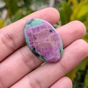 May include: An oval-shaped cabochon gemstone featuring a vibrant magenta-pink centre with visible crystalline textures and natural white veining, surrounded by a dark forest green border containing small black mineral inclusions.