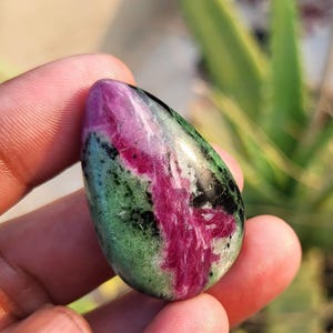 Könnte beinhalten: Ein polierter Edelstein in Tropfenform. Der Stein weist eine lebendige Mischung aus Farben auf, darunter tiefes Rot, Grün und Schwarz. Das Rot zieht sich über den Stein, während das Grün und Schwarz einen gesprenkelten Effekt erzeugen. Der Stein wird in einer Hand gehalten.