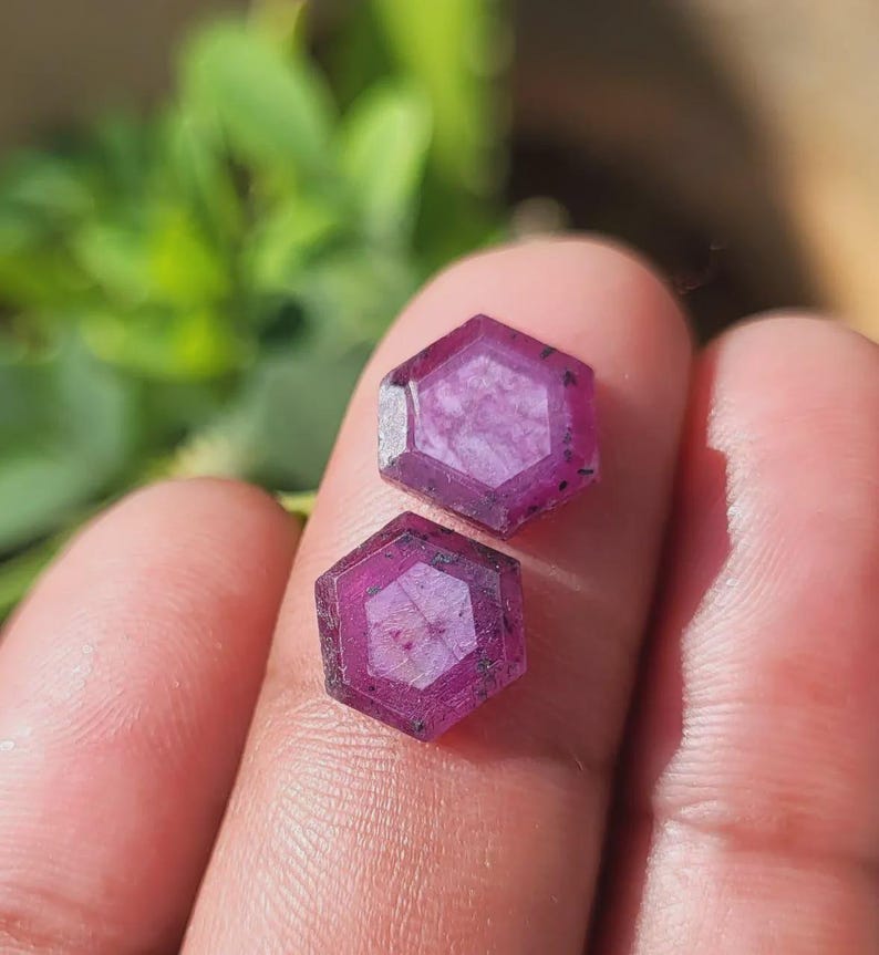 May include: Two hexagonal, faceted gemstones with a deep magenta hue. The gems are held between fingers, with a blurred green background. The gems have a complex internal structure and a polished surface, suitable for jewelry.
