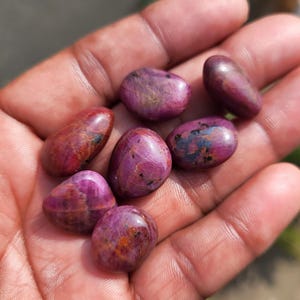 Könnte beinhalten: Eine Sammlung polierter Rubin-Edelsteine in verschiedenen Rot- und Rosatönen. Die Steine haben eine glatte, abgerundete Form und zeigen natürliche Muster und Einschlüsse. Die Edelsteine werden in einer offenen Hand gehalten, um ihre Farbe und Textur zu präsentieren.
