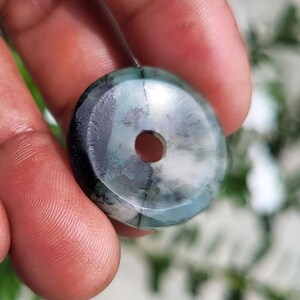 May include: A circular, donut-shaped pendant featuring a polished surface with a marbled pattern of deep forest green, charcoal gray, and creamy white tones. The piece has a smooth, rounded edge and a centered circular hole for threading.
